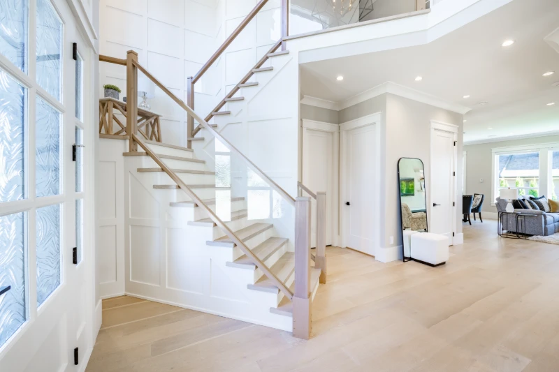 Bright, modern foyer with light wood floors, white walls, and a wooden staircase with glass panels. A large mirror and white bench sit by the stairs, and a living room is visible in the background.