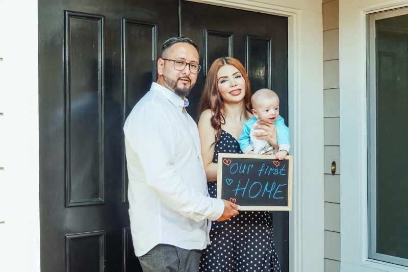 Young family with baby holding a sign Our First Home at door