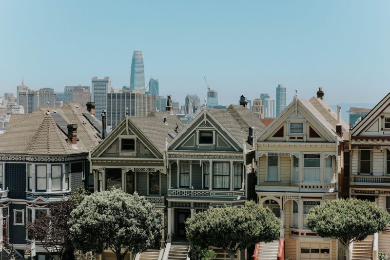 Colorful Victorian houses line a street with the San Francisco city skyline in the background.