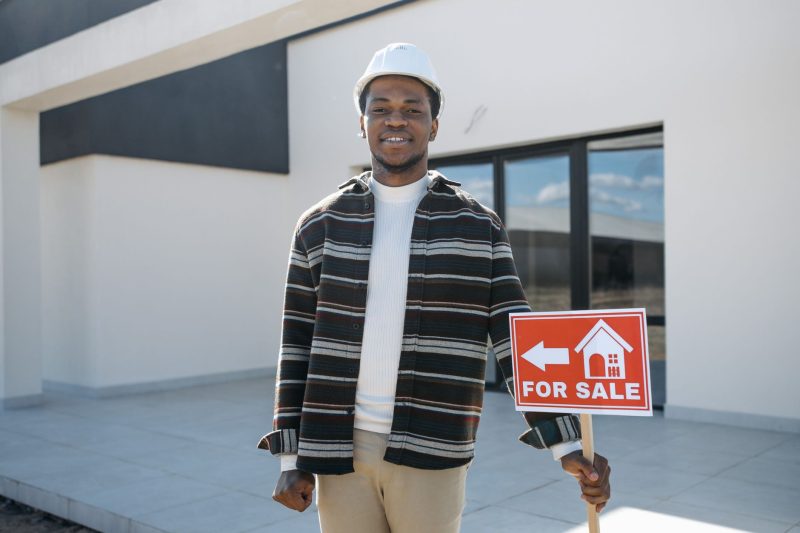 Young Black male realtor smiling while holding a for sale sign in front of a modern house.
