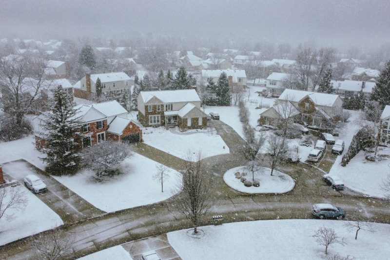 Aerial view of a snowy suburban neighborhood with lined houses and quiet streets under a clear sky.